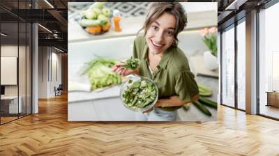 Portrait of a young and cheerful woman eating salad standing on the kitchen with food ingredients on the background, view from above. Vegetarianism, wellbeing and healthy lifestyle concept Wall mural