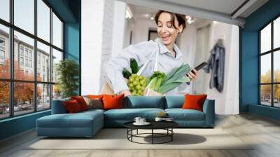 Portrait of a young and cheerful woman standing with mobile phone and shopping bag full of fresh food at home. Concept of buying products online Wall mural