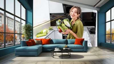 Portrait of a young and cheerful woman with healthy raw food on the kitchen at home. Vegetarianism, wellbeing and healthy lifestyle concept Wall mural