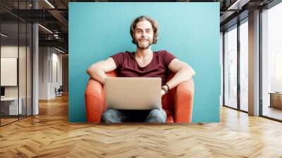 Portrait of a young caucasian bearded man with long hair dressed in t-shirt working with laptop on the chair on the colorful background Wall mural