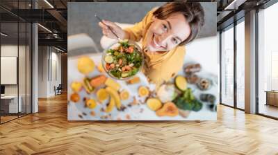 Portrait of a young cheerful woman eating salad at the table full of healthy raw vegetables and fruits on the kitchen at home, view from above. Concept of vegetarianism, healthy eating and wellness Wall mural