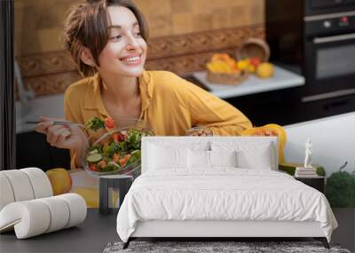 Portrait of a young cheerful woman eating salad at the table full of healthy raw vegetables and fruits on the kitchen at home. Concept of vegetarianism, healthy eating and wellness Wall mural