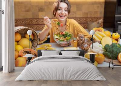 Portrait of a young cheerful woman eating salad at the table full of healthy raw vegetables and fruits on the kitchen at home. Concept of vegetarianism, healthy eating and wellness Wall mural