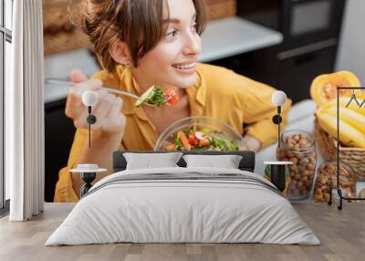 Portrait of a young cheerful woman eating salad at the table full of healthy raw vegetables and fruits on the kitchen at home. Concept of vegetarianism, healthy eating and wellness Wall mural