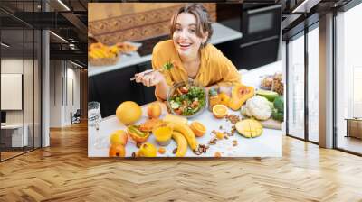 Portrait of a young cheerful woman eating salad at the table full of healthy raw vegetables and fruits on the kitchen at home. Concept of vegetarianism, healthy eating and wellness Wall mural