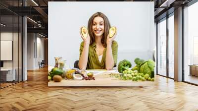 Portrait of a young cheerful woman with sliced avocado and lots of healthy green food on the table. Concept of vegetarianism and well-being Wall mural