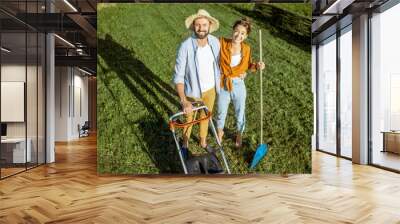 Portrait of a young couple standing together on the green lawn while cleaning backyard with lawn mower and rakes Wall mural