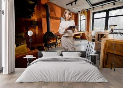 Portrait of a young pretty woman standing with a digital tablet in cozy living room. Wide view on interior in loft style with burning fireplace on background Wall mural