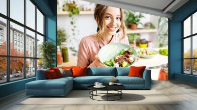 Portrait of a young woman holding a plate of salad sitting indoors surrounded with green flowers and healthy vegan food Wall mural