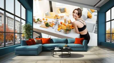 Portrait of an athletic woman having a break, drinking juice while standing with lots of healthy fresh food on the kitchen. Concept of losing weight, sports and healthy eating Wall mural