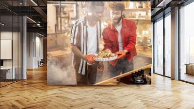 Portrait of two lovely multiracial guys standing together with cooked turkey on kitchen with a steam on background from the oven. Concept of gay relations everyday life together at home Wall mural