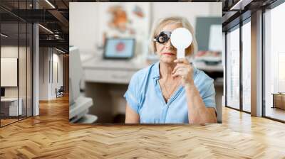 Senior woman checking vision with eye test glasses and scapula during a medical examination at the ophthalmological office Wall mural