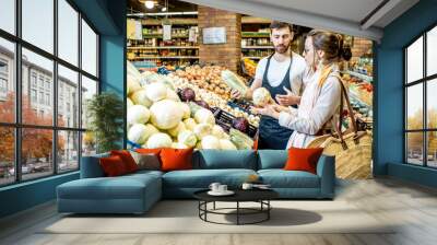 Shop worker helping young woman client to chooose vegetables in the supermarket Wall mural