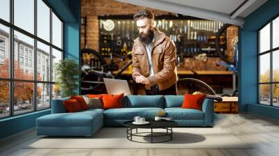 Stylish bearded businessman as a shop owner or manager working with laptop at the bicycle workshop Wall mural