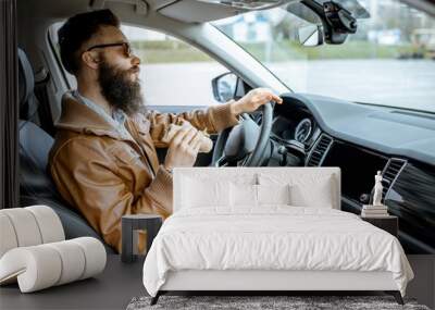 Stylish bearded man having a snack with tasty sandwich while driving a car in the city Wall mural