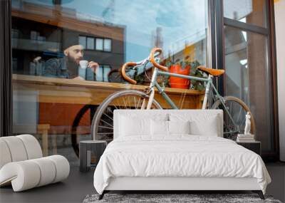 Stylish man enjoying coffee drink while sitting at the cafe near the window with retro bicycle. Wide view through the window Wall mural