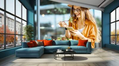 Stylish young woman eating healthy salad on a restaurant terrace, feeling happy on a summer day Wall mural