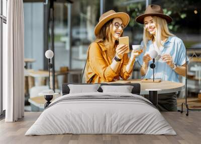 Two female best friends spending time together on the cafe terrace, feeling happy standing with coffee and phone during a summer day Wall mural