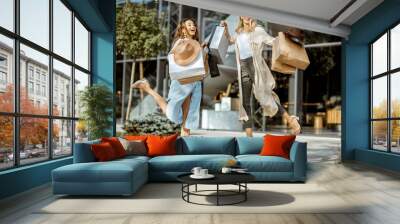 Two happy girlfriends jumpinh with shopping bags in front of the shopping mall outdoors, feeling excited with new purchases Wall mural