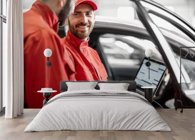 Two male auto mechanics in red uniform diagnosing car with computer standing near the luxury car at the workshop Wall mural