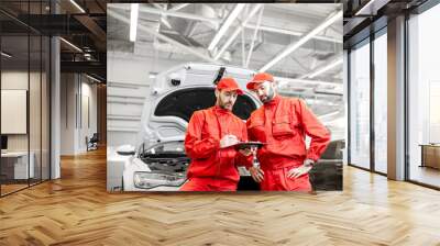 Two male auto mechanics in red uniform signing vehicle service documents standing near the car at the car service Wall mural