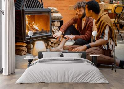 Two men talking while sitting together by the burning fireplace at cozy home. Concept of homosexual relations and coziness on winter time. Idea of multinational gay families Wall mural