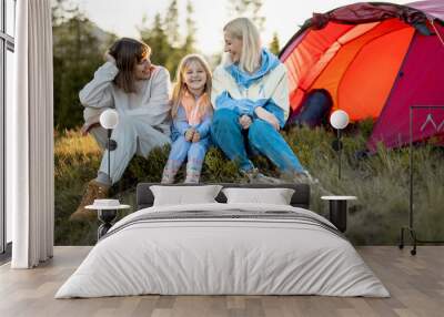 Two sisters or friends sit together with a little girl and have lovely conversation at campsite, sitting happily near tent on sunset. Homosexual family with a kid travel in mountains Wall mural