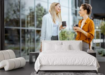 Two young businesswomen talking near the office building, having a small talk during the coffee break outdoors. Wall mural