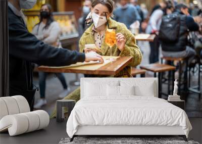 Waiter brings a menu for a young female customer at outdoor cafe. Woman wearing medical mask visiting public place Wall mural