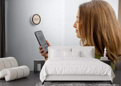 Woman dressed in green sweater regulating heating temperature with a modern wireless thermostat and smart phone at home. Synchronization of thermostat with mobile devices concept Wall mural