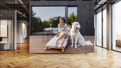 Woman enjoys the nature while sits on sunbed on wooden terrace at the modern hotel resort with panoramic windows near pine forest whith big white dog. Concept of solitude and recreation on nature Wall mural