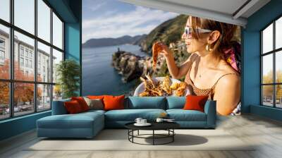 Woman holds plate with seafood at the coast with beautiful landscape of Vernazza village in Italy. Mediterranean food concept. Idea of traveling famous Cinque Terre region Wall mural