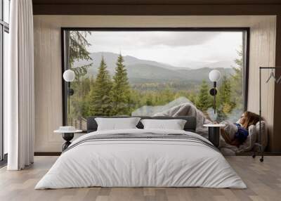 Woman lying on window sill covered with blanket, enjoying great view on mountains and pine forest during vacation in house on nature Wall mural