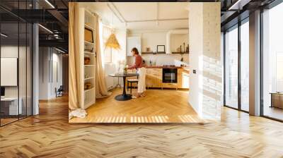 Woman puts flowers on a dinner table, decoration and having some house chores at home. Wide interior view on sunny studio apartment in beige tones Wall mural