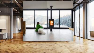 Woman sitting with dog near fireplace and panoramic window at modern living room with stunning view on snowy mountains. Concept of rest in houses or cabins on nature. Idea of escape from everyday life Wall mural