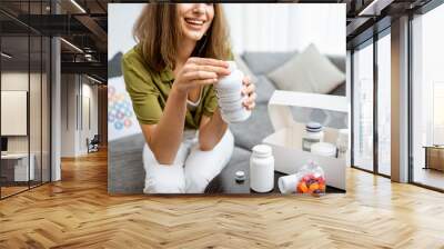 Woman taking vitamins or nutritional supplements in the form of pills while sitting on the couch at home. Concept of biohacking and preventive medicine Wall mural