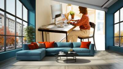 Woman works on computers while sitting by wooden table at sunny room at home. Concept of remote work from home at cozy atmosphere Wall mural