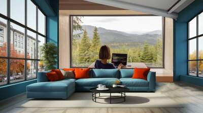 Woman works on laptop while sitting by the table in front of panoramic window with great view on mountains. Remote work and escaping to nature concept Wall mural