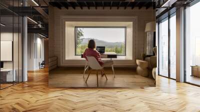 Woman works on laptop while sitting by the table in front of panoramic window with great view on mountains. Wide interior view. Remote work and escaping to nature concept Wall mural