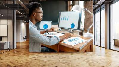 Young african man working with analitycs sitting at the beautiful working place with computer, laptop and documents Wall mural