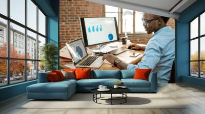 Young african man working with analitycs sitting at the beautiful working place with computer, laptop and documents Wall mural