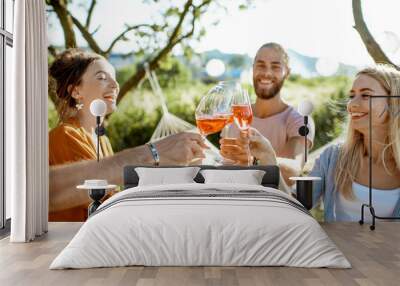 Young and cheerful friends having fun, clinking with wine glasses in the beautifully decorated backyard during a festive meeting or party on a sunny summer evening Wall mural