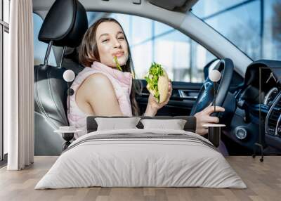 Young and cheerful woman in sportswear eating healthy sandwich with salad while driving car in the city Wall mural