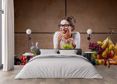 young and cute woman eating grapes at the table full of fruits and vegetables in the wooden interior Wall mural