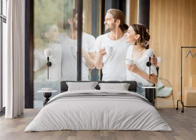 Young and happy couple dressed in white shirts standing together with cups near the window at home Wall mural