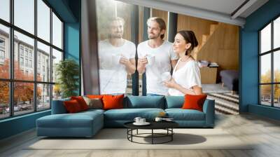 Young and happy couple dressed in white shirts standing together with cups near the window at home Wall mural