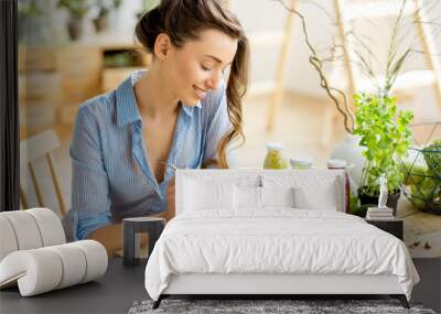 young and happy woman eating healthy salad sitting on the table with green fresh ingredients indoors Wall mural