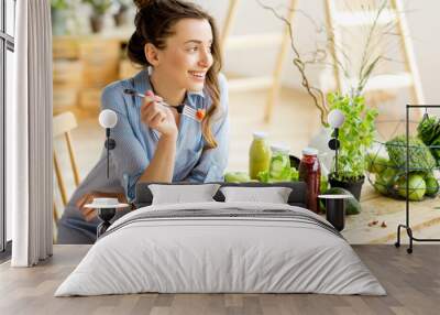 Young and happy woman eating healthy salad sitting on the table with green fresh ingredients indoors Wall mural