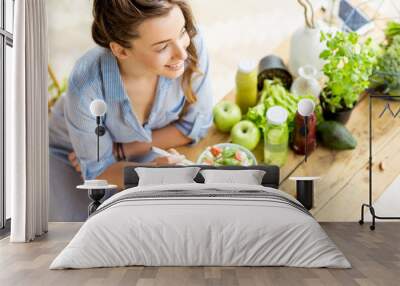 young and happy woman eating healthy salad sitting on the table with green fresh ingredients indoors Wall mural