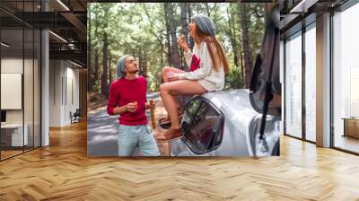 Young and lovely couple in sweaters and hats having fun, eating baguette with jam near the car on the roadside in the pine forest. Young family having quick snack while traveling Wall mural